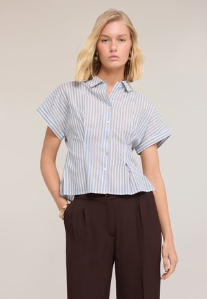 Blonde vrouw die een blauw-wit gestreept, kortemouwen, knoophemd draagt en donkerbruine, hooggetailleerde broek met hand in zak.