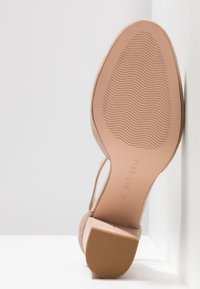 Chaussure à talon haut beige avec une texture douce semblable au daim et une semelle en caoutchouc. Les caractéristiques notables incluent un talon sculpté et un motif de grip ondulé sur la semelle.