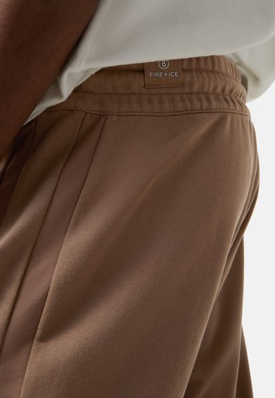 Brune træningsbukser med en struktureret stof, der har en kontrasterende sidelapel, elastisk talje og en syet brandlabel på bagsiden.