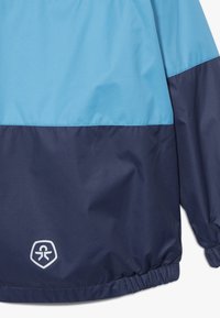 Modrá a námořnická větrovka s hladkou nylonovou texturou, elastickými manžetami a emblémem na dolní levé straně.