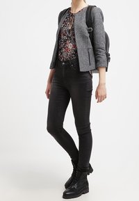 Blazer grigio con texture sopra una blusa floreale, abbinato a jeans skinny neri e stivali neri con lacci. Zaino nero indossato sulle spalle.