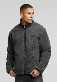 Gray fleece jacket with a high collar, front zipper, and side pockets; features a textured exterior and a black logo patch on the chest.