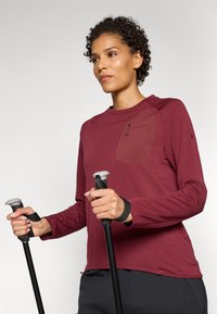 Chemise à manches longues bordeaux avec un panneau texturé et une poche zippée sur la poitrine. Fabrication avec un tissu extensible et des poignets ajustés.  Empêche les bâtons de randonnée.