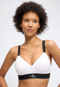 Γυναίκα με σγουρά μαλλιά που φοράει λευκό bralette Calvin Klein με μαύρα λουράκια και ζώνη, κοιτάζοντας ελαφρώς προς το πλάι.