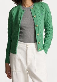 Grön kabelstickad cardigan med rund halsringning, sex stora knappar, ribbade ärmslut och en liten logodetalj. Bärs över en grå topp.