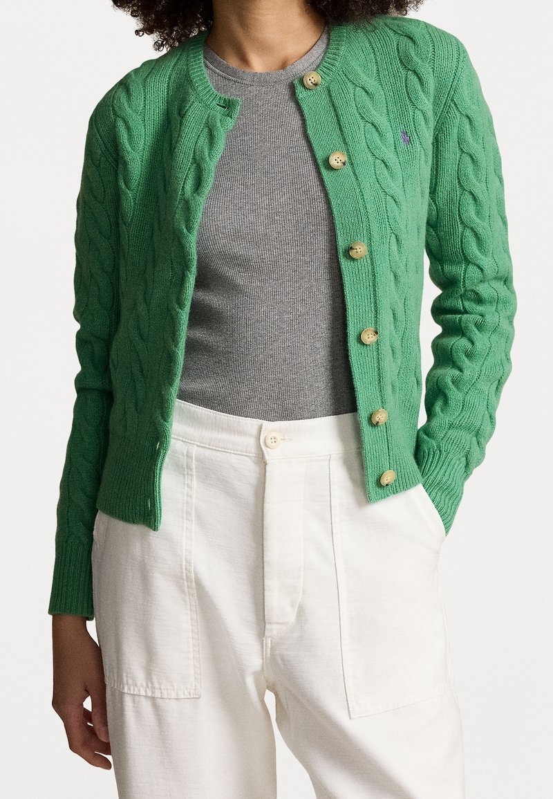 Grön kabelstickad cardigan med rund halsringning, sex stora knappar, ribbade ärmslut och en liten logodetalj. Bärs över en grå topp.