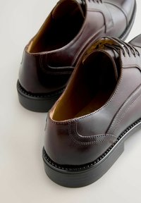 Zapatos de cuero marrón con una textura suave, punta redondeada y suela de goma negra. Presentan un diseño con cordones y detalles de costuras en contraste.