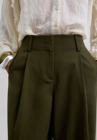 Mujer vistiendo pantalones de tiro alto en color verde oliva con bolsillos y una blusa abotonada arrugada de color crema, manos en los bolsillos, pulsera de plata y verde.