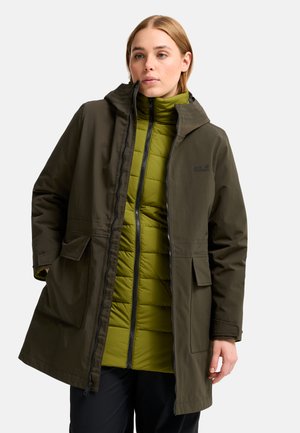 Donna che indossa una giacca imbottita verde sotto un cappotto aperto con cappuccio verde oliva scuro con tasche con pattina e pantaloni neri.