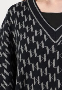 Sveter s výstrihom do V, ktorý má geometrický vzor v sivých odtieňoch na tmavom námořníckom pozadí, so žebrovanými detailmi pozdĺž výstrihu a spodného okraja.