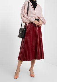 Roter Faltenrock mit glänzender Oberfläche, kombiniert mit einer hellrosa gestreiften Bluse und einer schwarzen Umhängetasche. Model trägt pfirsichfarbene Heels.