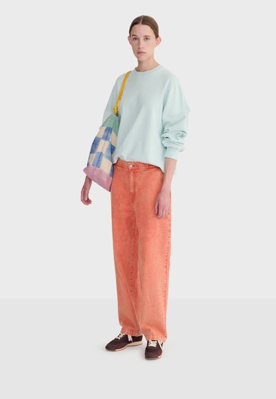 Mladá žena stojaca, oblečená v svetlomodrej mikine, voľných oranžových nohaviciach, tmavých teniskách a držiaca pastelovú kockovanú nákupnú tašku so žltými rúčkami.