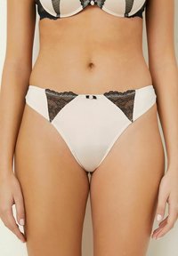 Béžové vysoké kalhotky s černými krajkovými panely a malou mašličkou s růžovým akcentem uprostřed na bedrech ženy.