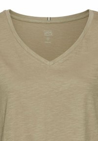 Beżowy organiczny t-shirt z dekoltem w kształcie litery V, o teksturowanym wykończeniu i subtelnym połysku, z metką wskazującą rozmiar (S) oraz materiał.