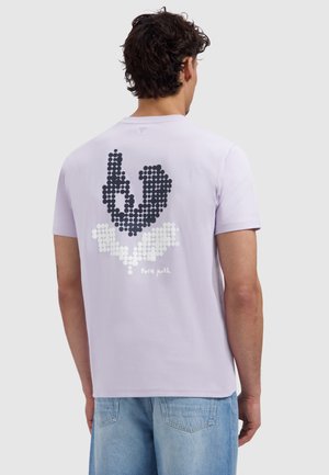 Man draagt een lichtpaarse T-shirt met een zwart-wit stippenpatroon dat abstracte vormen vormt, en lichtblauwe spijkerbroek.