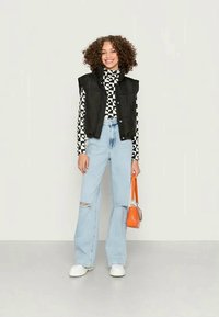 Colete acolchoado preto sobre uma camisola de gola alta com padrão preto e branco, combinado com jeans rasgados azul claro e sapatos brancos, segurando uma bolsa laranja.