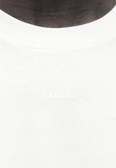 Gros plan sur une personne portant un t-shirt blanc à col rond avec le texte embossé "SANDRO" centré sur la poitrine.
