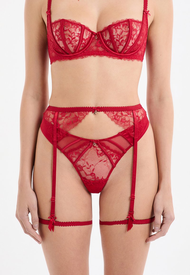 Vrouw in een rood kanten lingeriesetje met beha, jarretel en bijpassende slip voor een witte achtergrond.