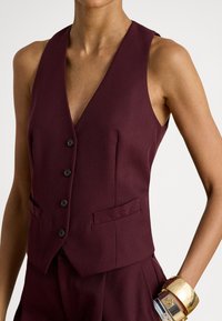Μπορντό ραμμένο γιλέκο με V-λαιμόκοψη, πέντε κουμπιά και πλαϊνές τσέπες. Κατασκευασμένο από υφή ύφασμα, συνοδευόμενο από χρυσά βραχιόλια στον καρπό.