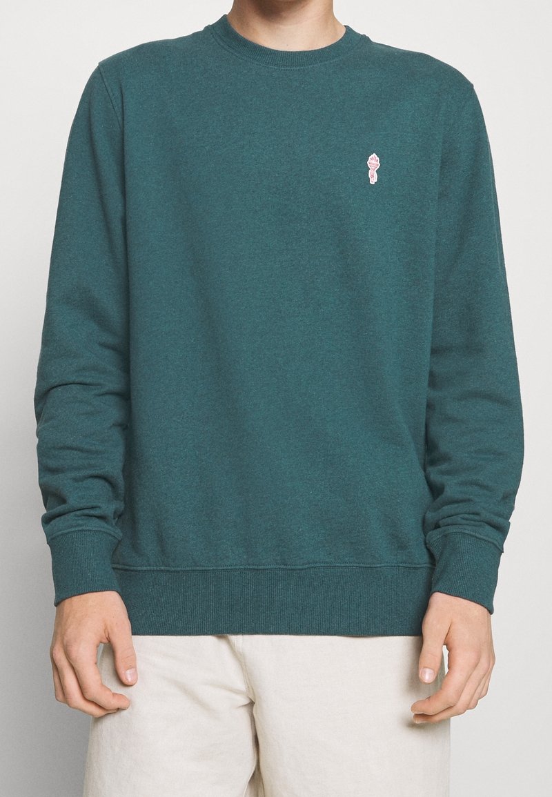 Homme portant un sweat-shirt col rond bleu sarcelle avec un petit emblème de torche rose sur le côté gauche de la poitrine et un pantalon beige clair, debout devant un fond blanc.