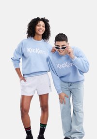 Felpe di colore azzurro chiaro con logo "Kickers", pantaloni corti grigi e jeans in denim. Gli accessori includono occhiali da sole e calze a fantasia.
