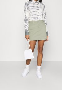 Donna che indossa una camicia grigia e bianca con stampa tie-dye, una mini gonnellina verde chiaro, sneaker bianche e tiene in mano una piccola borsa bianca.