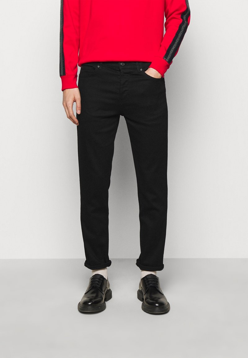 Personne portant un pantalon noir ajusté, des chaussures habillées noires, des chaussettes blanches et une chemise rouge à manches longues avec des rayures noires sur les manches, debout contre un mur blanc.