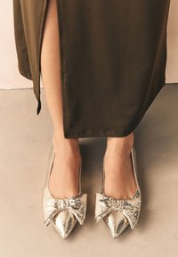 Auksinės gyvatės odos plokšti batai su dideliu kaspinu priekyje. Batai yra smailūs ir turi lygią, blizgančią apdailą.