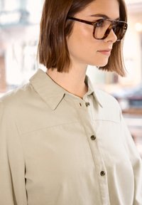 Femme aux cheveux courts bruns portant des lunettes à monture écaille de tortue et une chemise beige boutonnée, regardant vers la droite dans une pièce à la lumière douce.