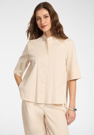 Kobieta w luźnej beżowej koszuli zapinanej na guziki z rękawami do łokcia i pasujących spodniach, stoi z jedną ręką w kieszeni.