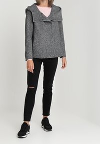 Šedá mikina s kapucí a klopovým límcem, se dvěma předními kapsami, nosena přes světle růžový top, v kombinaci s černými skinny džíny a černými teniskami.