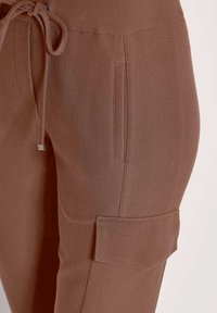 Braune texturierte Hose mit seitlicher Klappentasche und Kordelzugbund, Nahaufnahme im oberen Oberschenkelbereich.