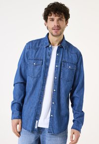 Blauwe denimshirt met knoopsluiting aan de voorkant, twee borstzakken en lange mouwen. Draagt over een wit T-shirt, met een casual pasvorm en een zachte textuur.