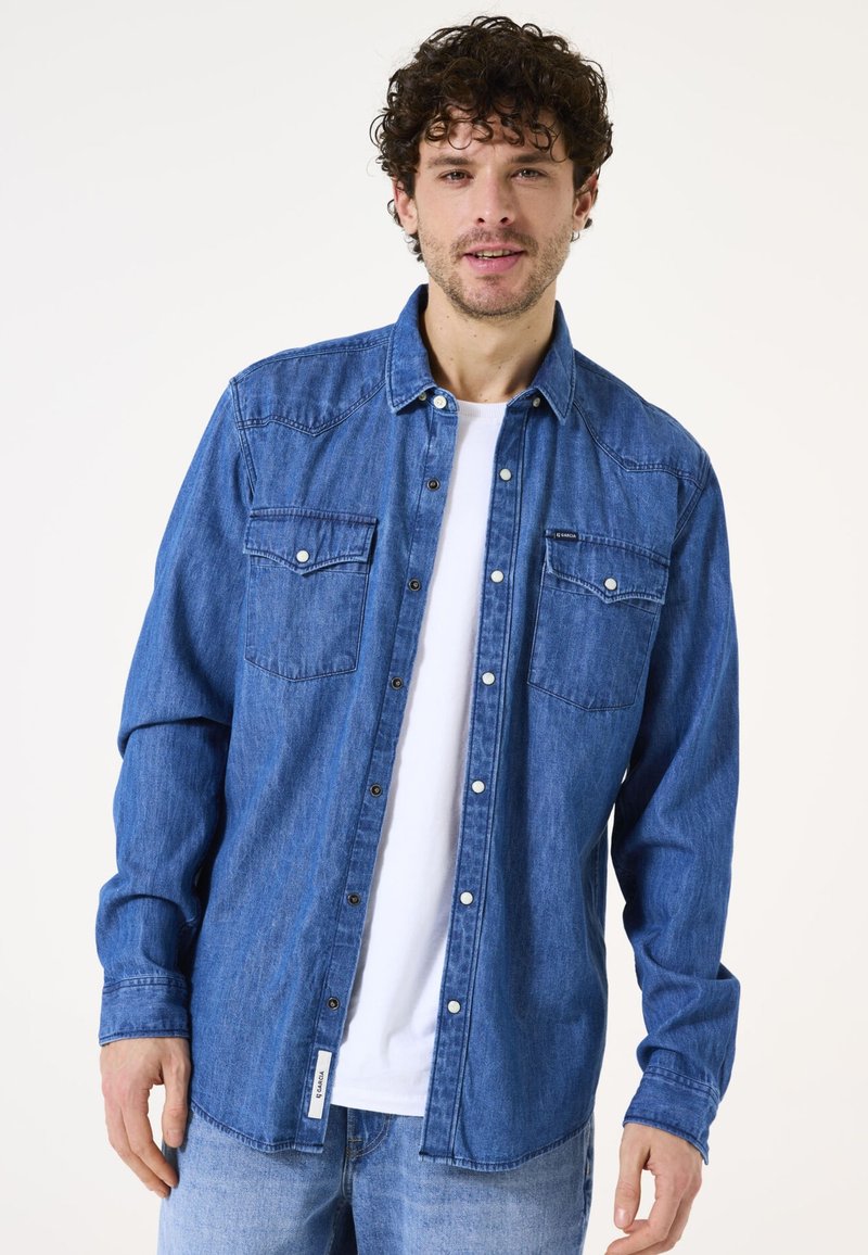 Blauwe denimshirt met knoopsluiting aan de voorkant, twee borstzakken en lange mouwen. Draagt over een wit T-shirt, met een casual pasvorm en een zachte textuur.