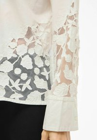 Balts blūze ar izšūtiem ziedu rakstiem caurspīdīgā audumā pie piedurknēm, ar vienkāršu aproci un melnu teksturētu fonu.