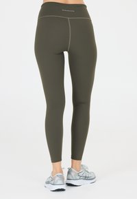 Olijfgroene leggings met een hoge tailleband, een gladde textuur en contrasterende stiksels. Gemaakt met reflecterende zilveren sneakers.
