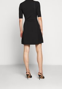 Femme debout, de dos, portant une robe noire mi-longue et des chaussures à talons hauts noires avec bride arrière sur un fond blanc.