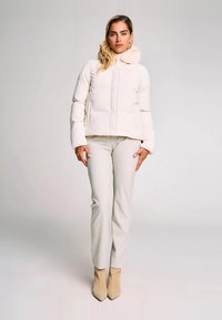 Donna che indossa una giacca a vento bianca, pantaloni chiari e stivali alla caviglia beige, in piedi contro uno sfondo grigio chiaro uniforme.