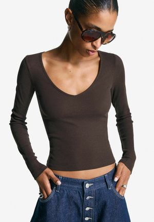 Femme portant de grandes lunettes de soleil écaille de tortue, un haut marron foncé à manches longues et col en V, et une jupe en jean taille haute boutonnée devant.