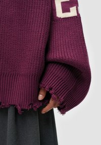 Übergroßer lila Strickpullover mit einem strukturierten Rippmuster, versehen mit ausgefransten Bündchen und Saum. Akzentuiert mit einem cremefarbenen Blockbuchstaben-Design.