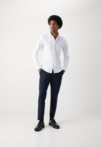 Hvit skjorte med lange ermer, kombinert med marineblå stripete bukser og svarte snøresko. Slim-fit silhuett. Glatt stoff.