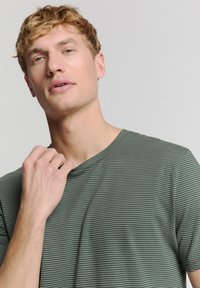 Groene t-shirt met korte mouwen en witte horizontale strepen, met een ruime halslijn en een zachte stof, gedragen door een persoon met licht haar.