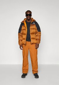 Casaco cria-marcas em castanho e preto, com fecho de correr e punhos elásticos, combinado com calças laranja. O modelo usa óculos de sol e sapatos pretos.