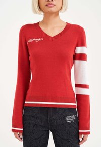 Roter gestrickter Pullover mit V-Ausschnitt, weißen Streifen an den Ärmeln, gerippten Bündchen und einem auf der Brust gestickten Logo.