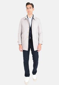 Cappotto beige chiaro con bottoni, pantaloni blu navy e scarpe da ginnastica bianche. Il cappotto presenta un taglio dritto e maniche lunghe, con una texture liscia.