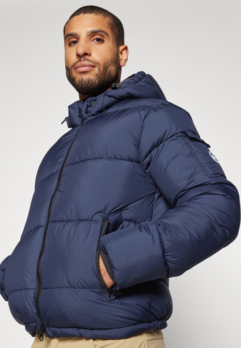 Mann mit kurz rasiertem Bart, der eine navyblaue Steppjacke mit Kapuze, die Hände in den Taschen, und beige Hosen trägt, leicht nach links gewandt vor einem schlichten Hintergrund.
