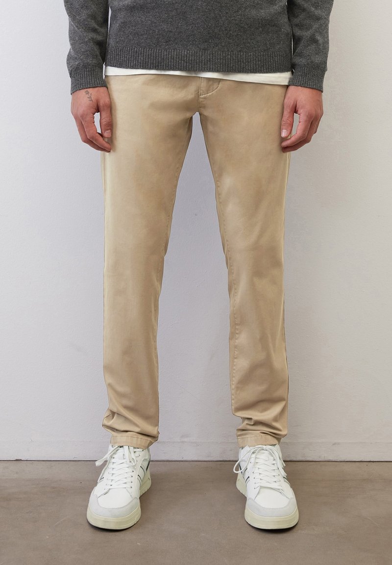 Personne portant un pantalon beige, des baskets blanches, un pull gris et une chemise blanche, debout devant un mur et un sol clairs et unis.