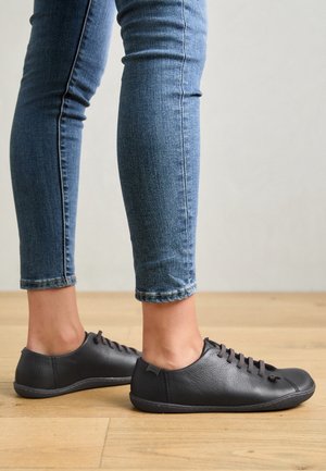 Jambes portant un jean bleu slim et des chaussures décontractées en cuir noir à lacets, debout sur un sol en bois devant un mur uni.