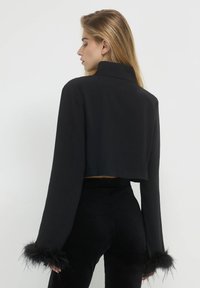 Schwarze cropped Jacke mit hohem Kragen, langen Ärmeln mit pelzigen Manschetten und glatter Textur, kombiniert mit schwarzen enganliegenden Hosen.