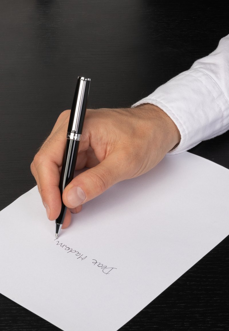 Main en chemise blanche écrivant "Chère Madame" avec un stylo noir sur une feuille blanche posée sur un bureau noir.